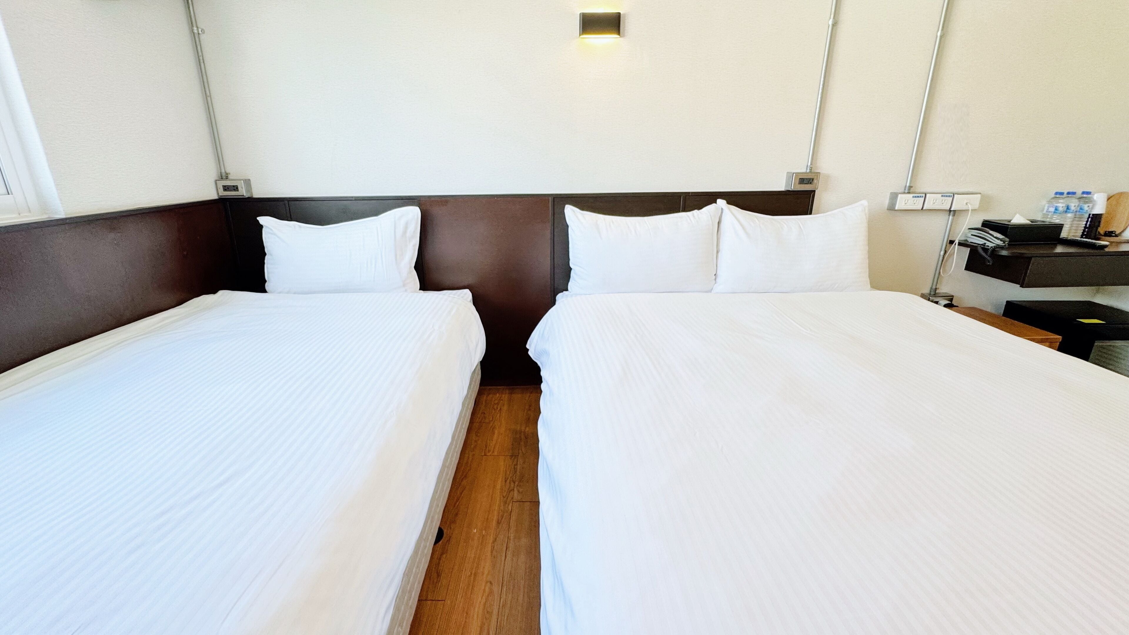 A hotel room with two beds, a wooden floor, a nightstand with a phone and a lamp, and a wall-mounted light fixture.