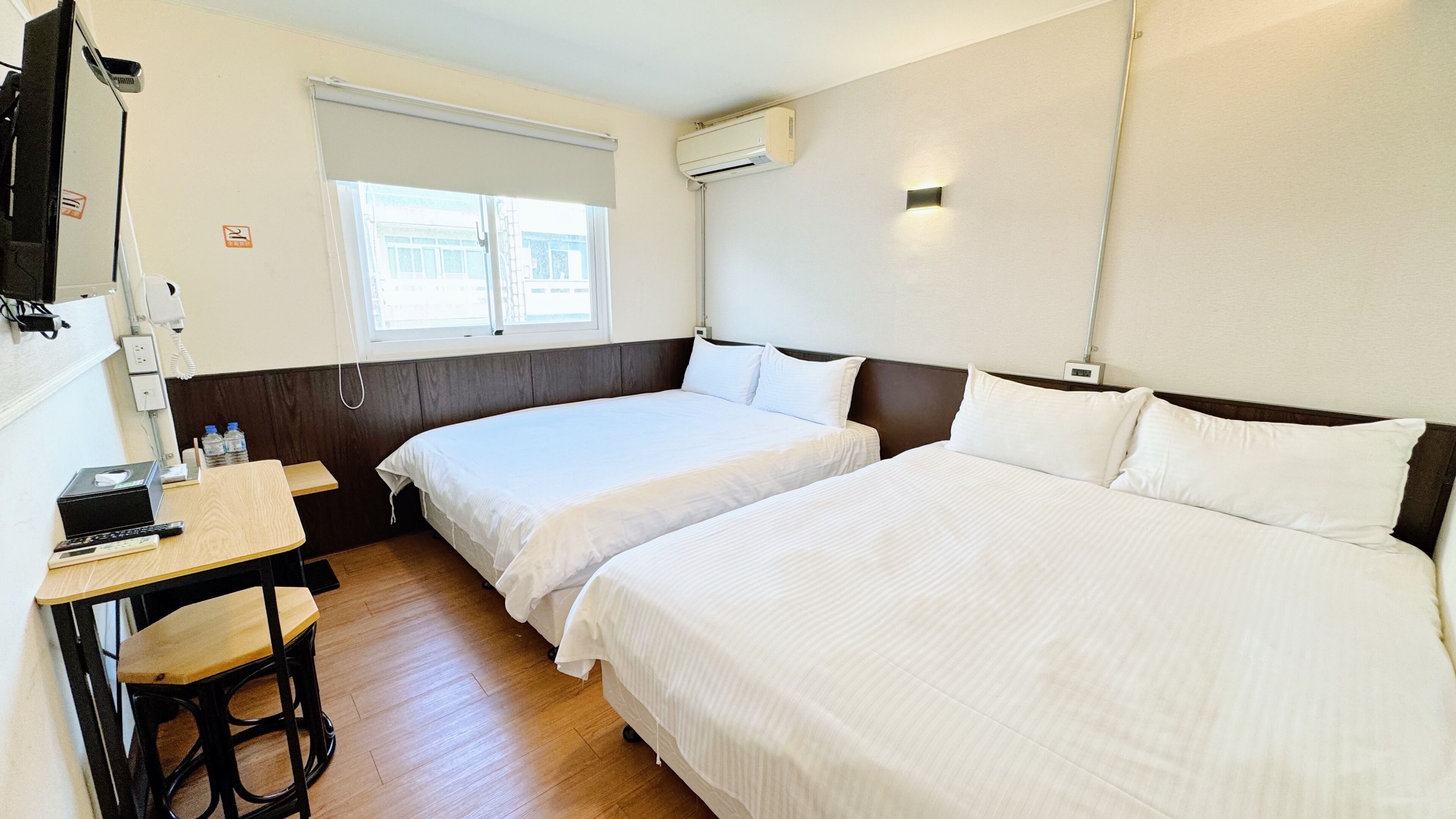 A hotel room with two beds, a desk with a chair, a telephone, and an air conditioning unit.
