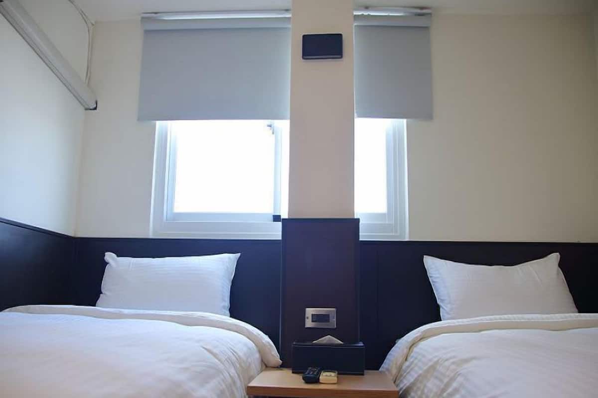 Habitación de hotel con dos camas, una mesita de noche, un termostato y una ventana con persianas.
