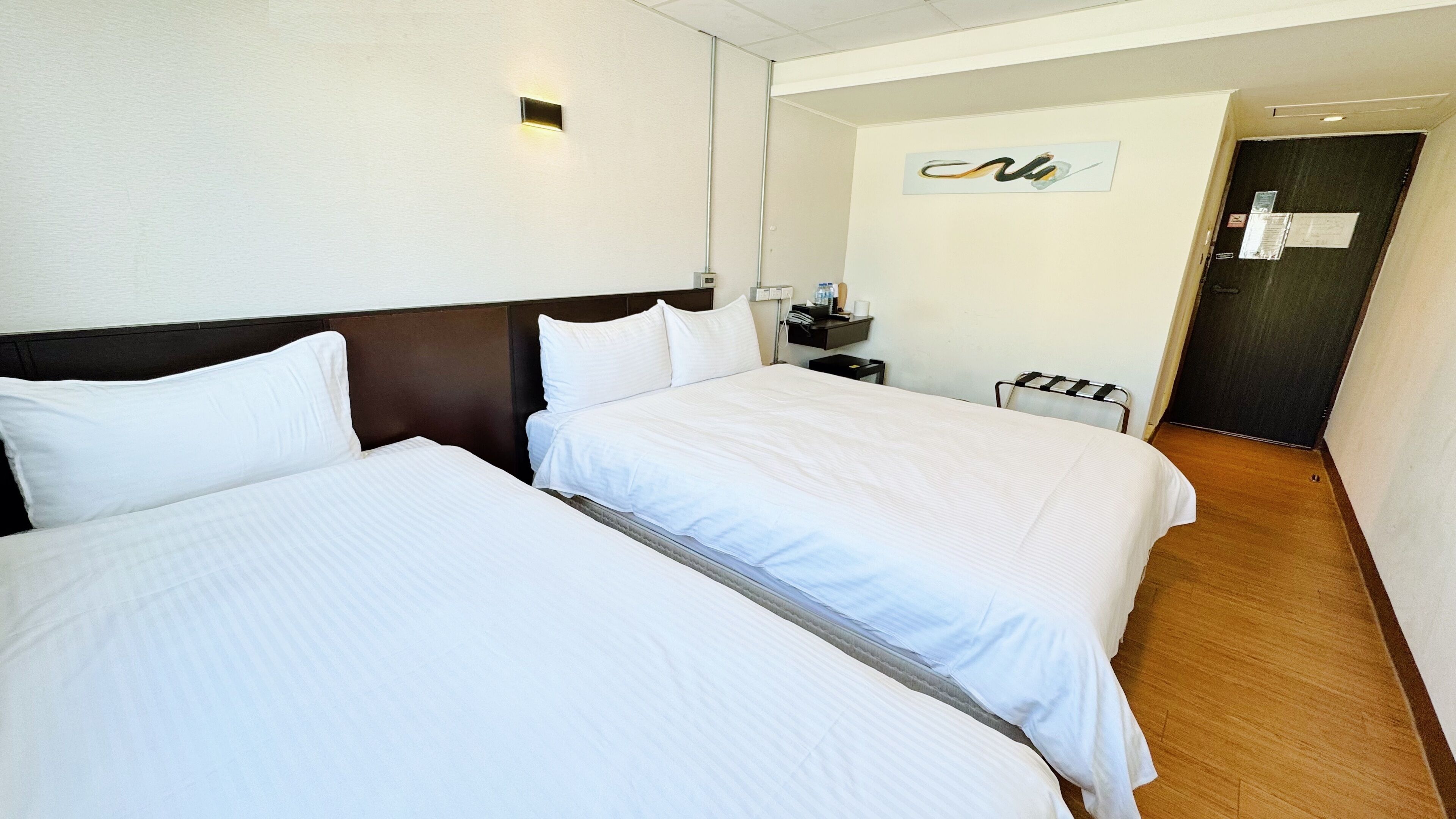 A hotel room with two beds, a nightstand, a wall-mounted light, and a door.