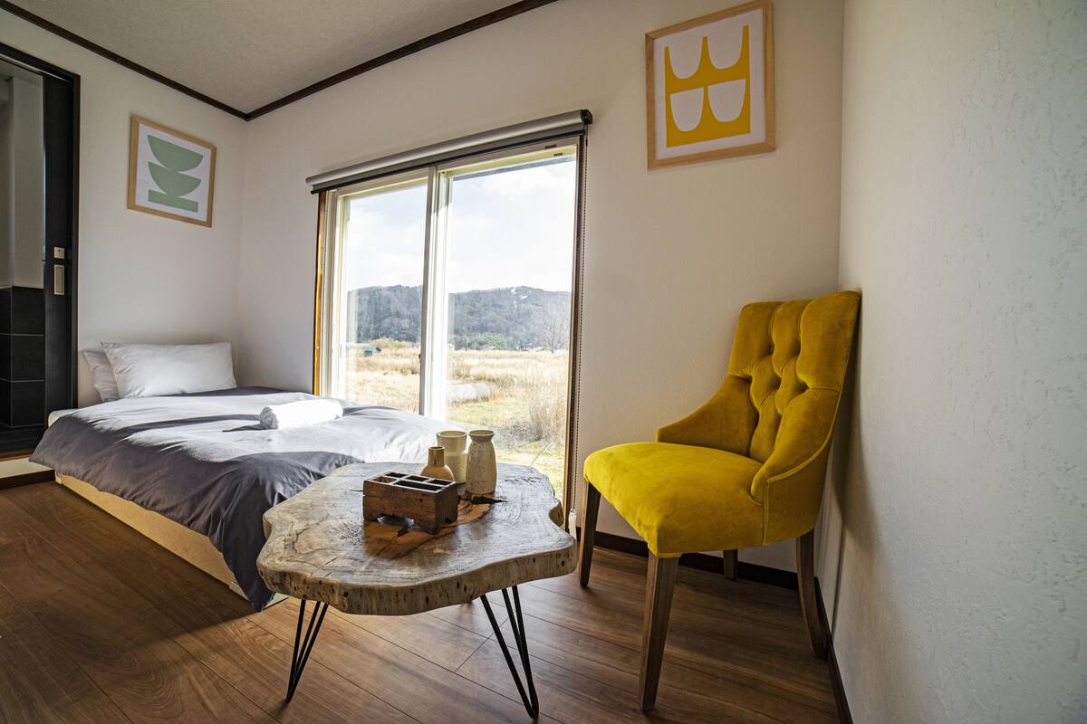 Une chambre Ă coucher avec un lit, un fauteuil jaune, une petite table avec un vase et une grande fenĂȘtre donnant sur lâextĂ©rieur.