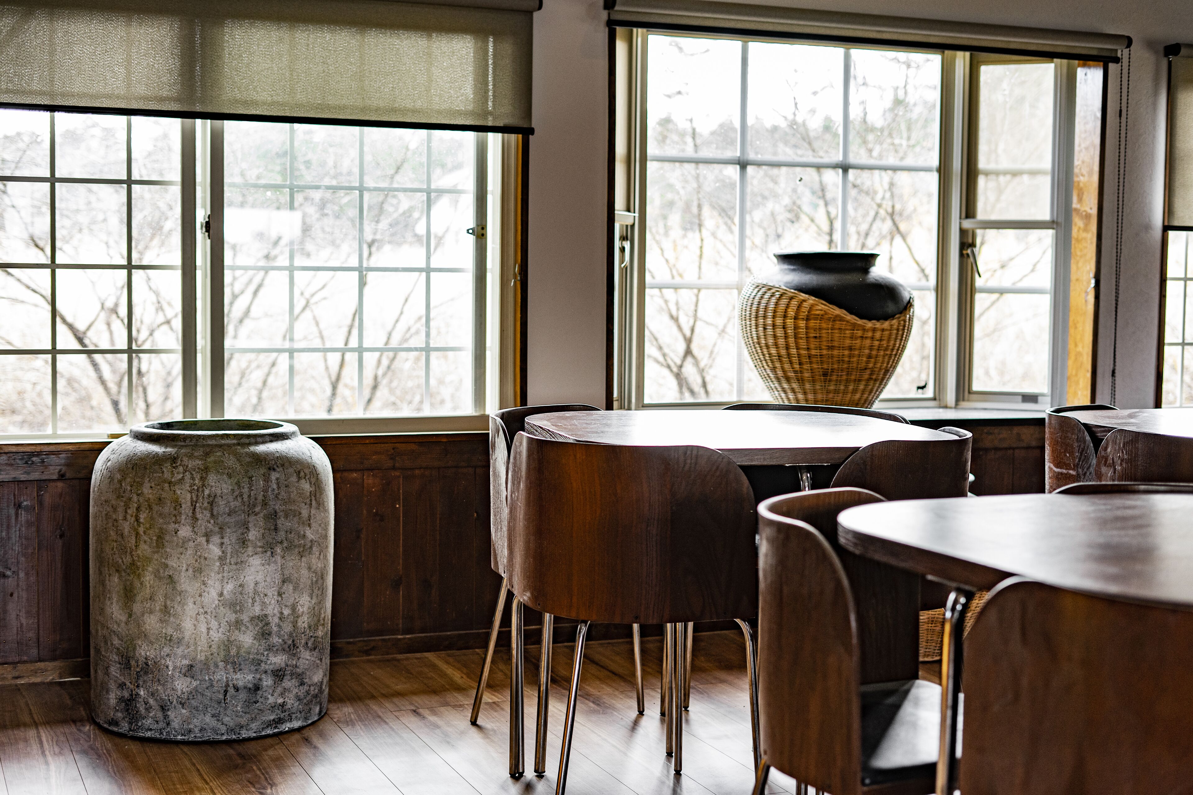 A dining area with wooden tables and chairs, large windows with a view of bare trees, and a large vase on one table.