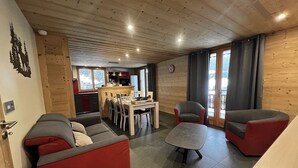 Een gezellige woonkamer met een rode bank, een grijze fauteuil en een eethoek met een houten tafel en stoelen.