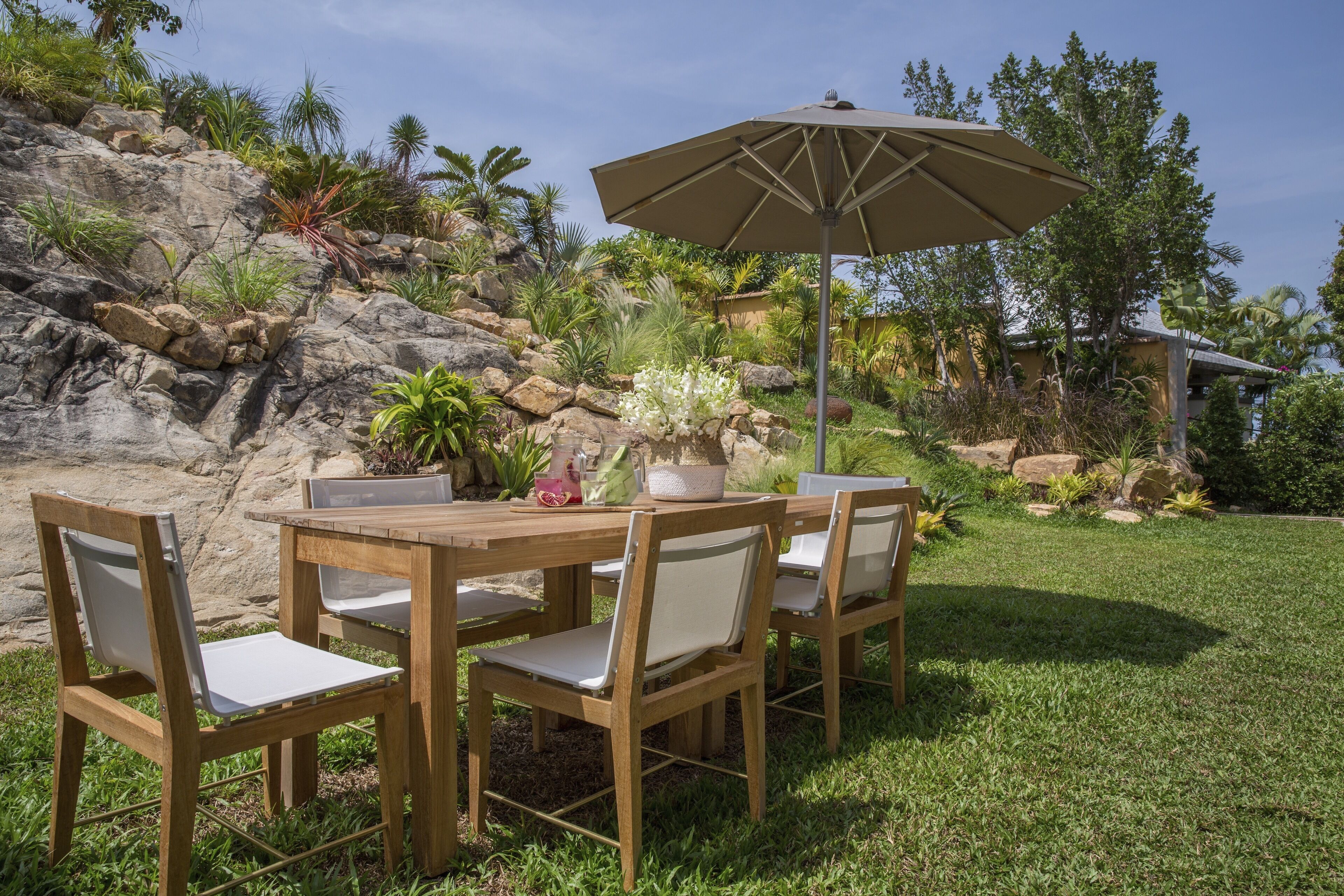 An outdoor dining area with a wooden table and chairs, an umbrella, and a garden with rocks and plants.