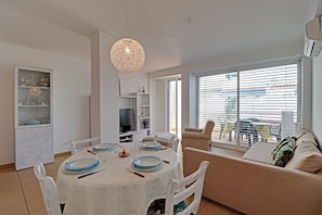 Ein Essbereich mit einem runden Tisch, Stühlen und einer Couch. Ein Fernseher und Regale mit Dekorationselementen sind ebenfalls vorhanden.