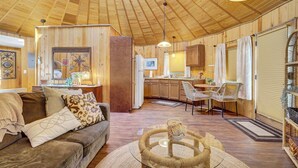 Interior acollidor d'una cabana amb sostre de fusta, zona de seients amb un sofà i una taula de cafè de vidre, cuina amb zona de dinar i una finestra gran amb cortines.