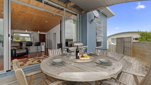 A wooden deck with a round table set for dining, a grill, and a sliding glass door leading to a living area with a sofa and a fireplace.