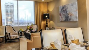 A hotel room with a view of a cityscape, a dining area with a table set for two, and a seating area with a sofa and armchairs.