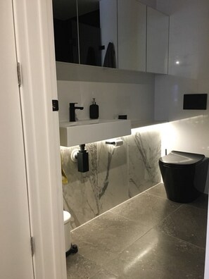 Un baño moderno con detalles de mármol en el revestimiento, inodoro negro y lavamanos blanco con dispensador de jabón.