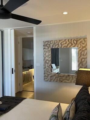 Un dormitorio con cama, ventilador de techo, un espejo grande con marco de madera y vista a la cocina contigua.