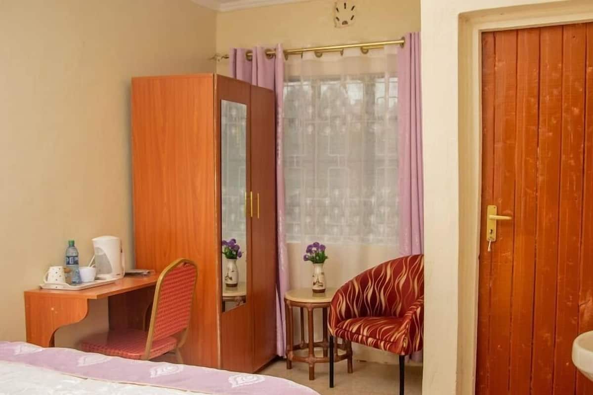 A hotel room with a bed, a desk with a chair, a small table with a chair, a wardrobe, and a window with curtains.