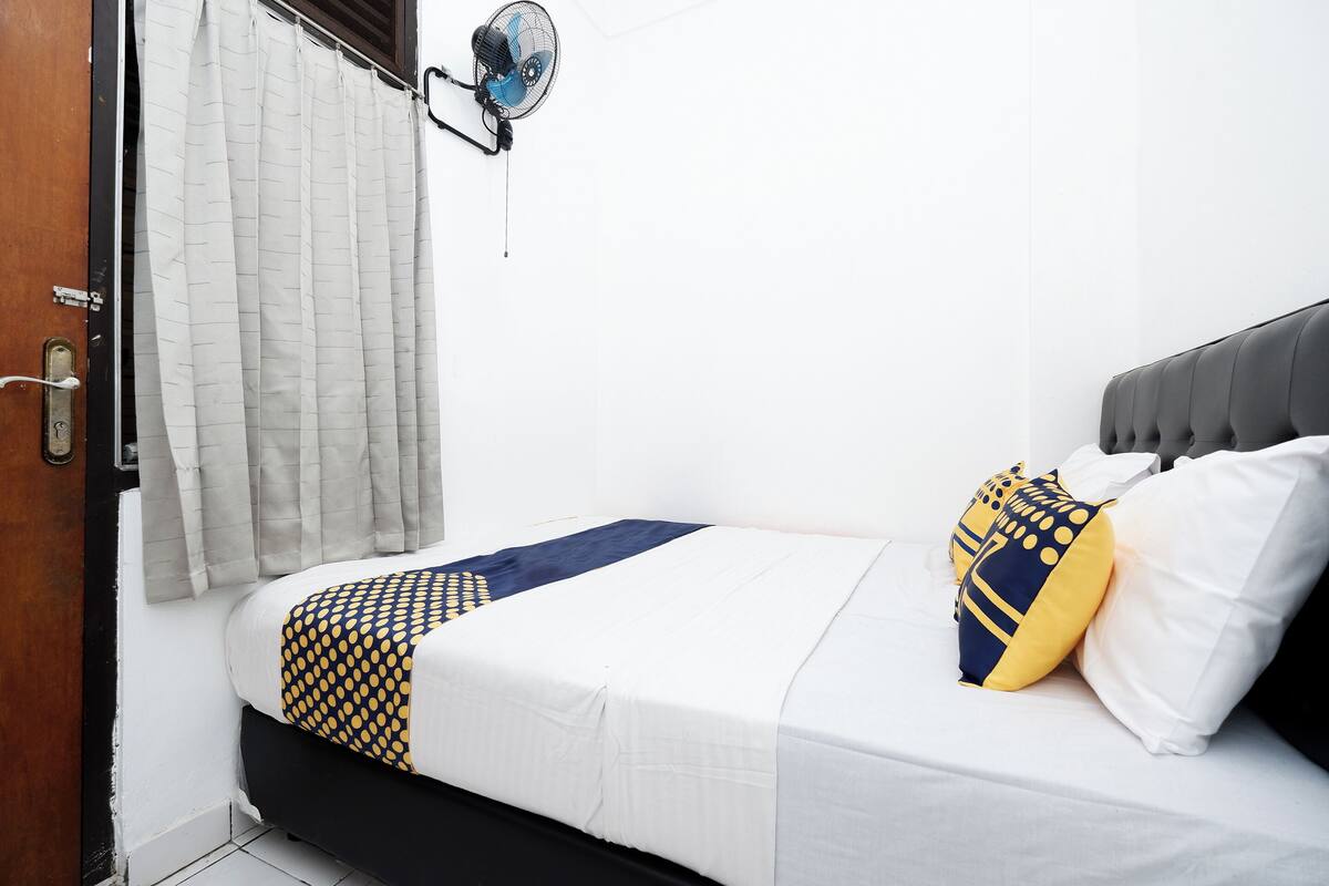 Una cama bien hecha con una colcha blanca y azul estampada, un cabecero tapizado en gris y dos almohadones con motivos amarillos y azules.