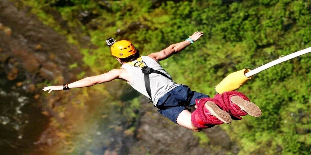 A person bungee jumping with a harness and helmet, suspended over a lush green forest.