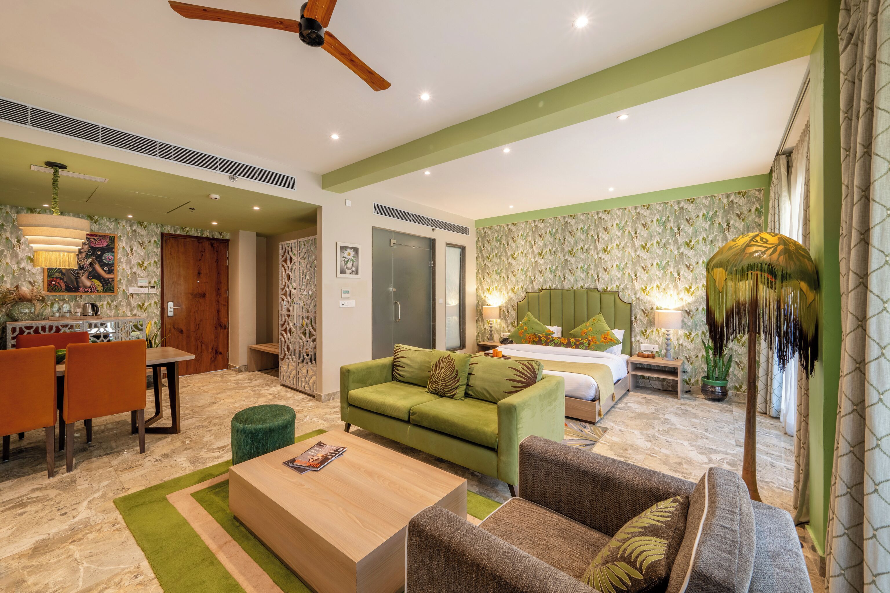 A hotel room with a green sofa, a wooden coffee table, a bed with a green headboard, and a dining area with a table and chairs.