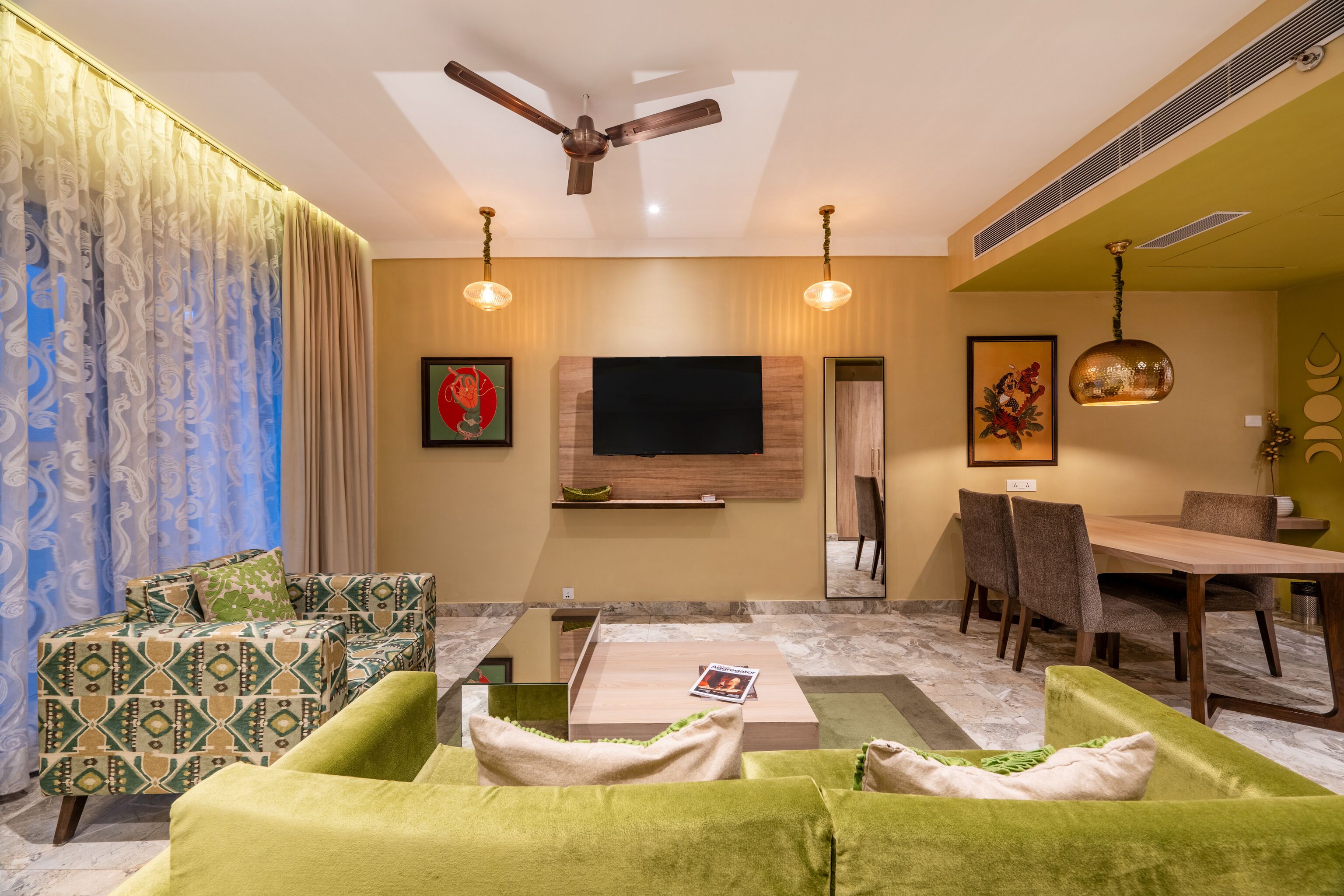 A modern living room with a flat-screen TV, green sofas, a coffee table, and a dining area with chairs and a table.