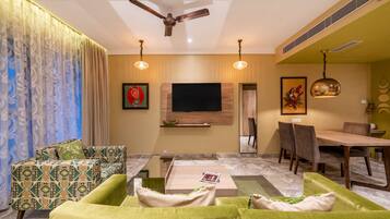 A modern living room with a flat-screen TV, green sofas, a coffee table, and a dining area with chairs and a table.