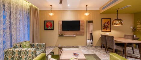 A modern living room with a flat-screen TV, green sofas, a coffee table, and a dining area with chairs and a table.