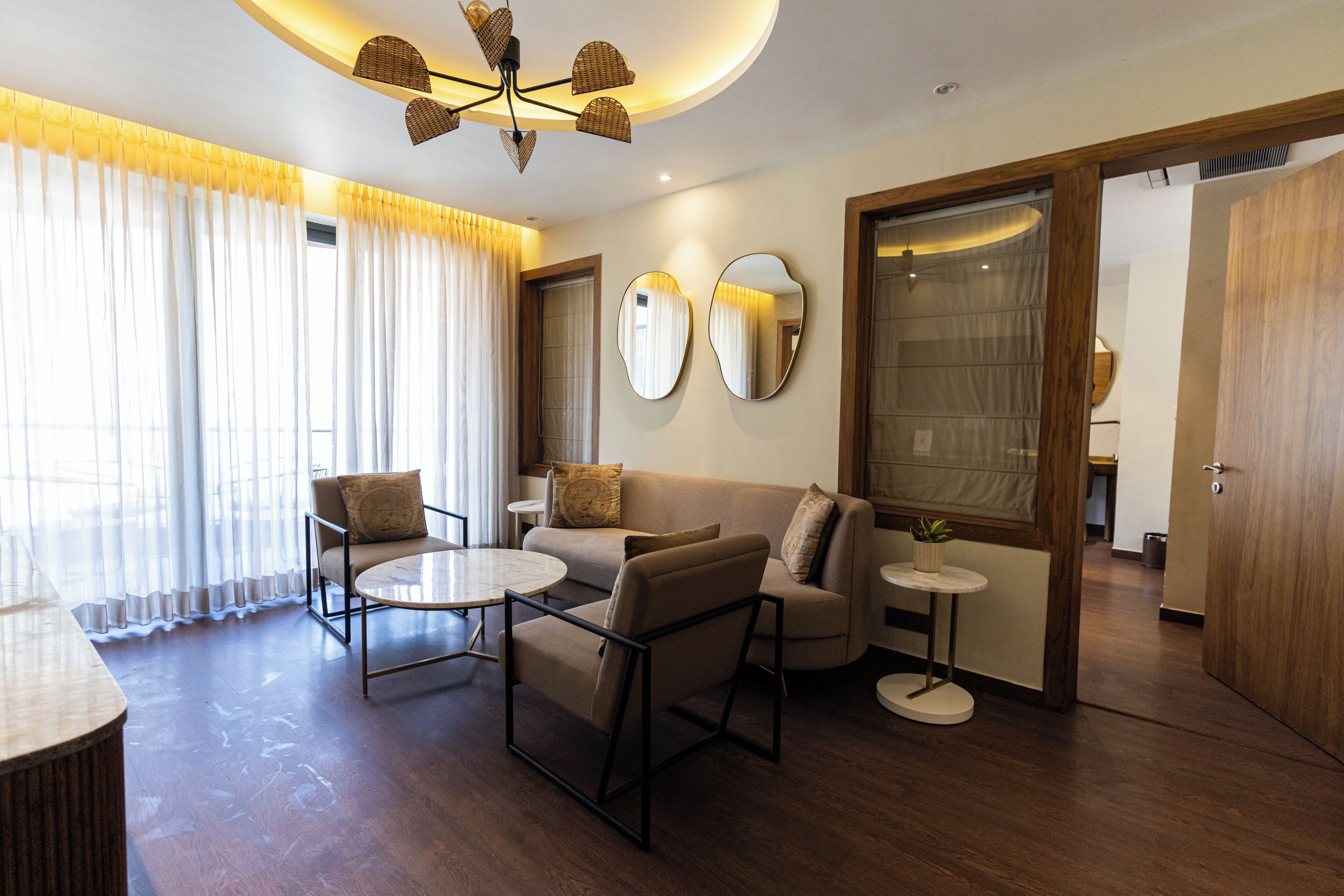 Un salon moderne comprenant un canapé, des fauteuils, une table basse et une petite table d’appoint. Deux miroirs sont accrochés au mur, ainsi qu’un luminaire au plafond et une porte qui donne sur une autre pièce.