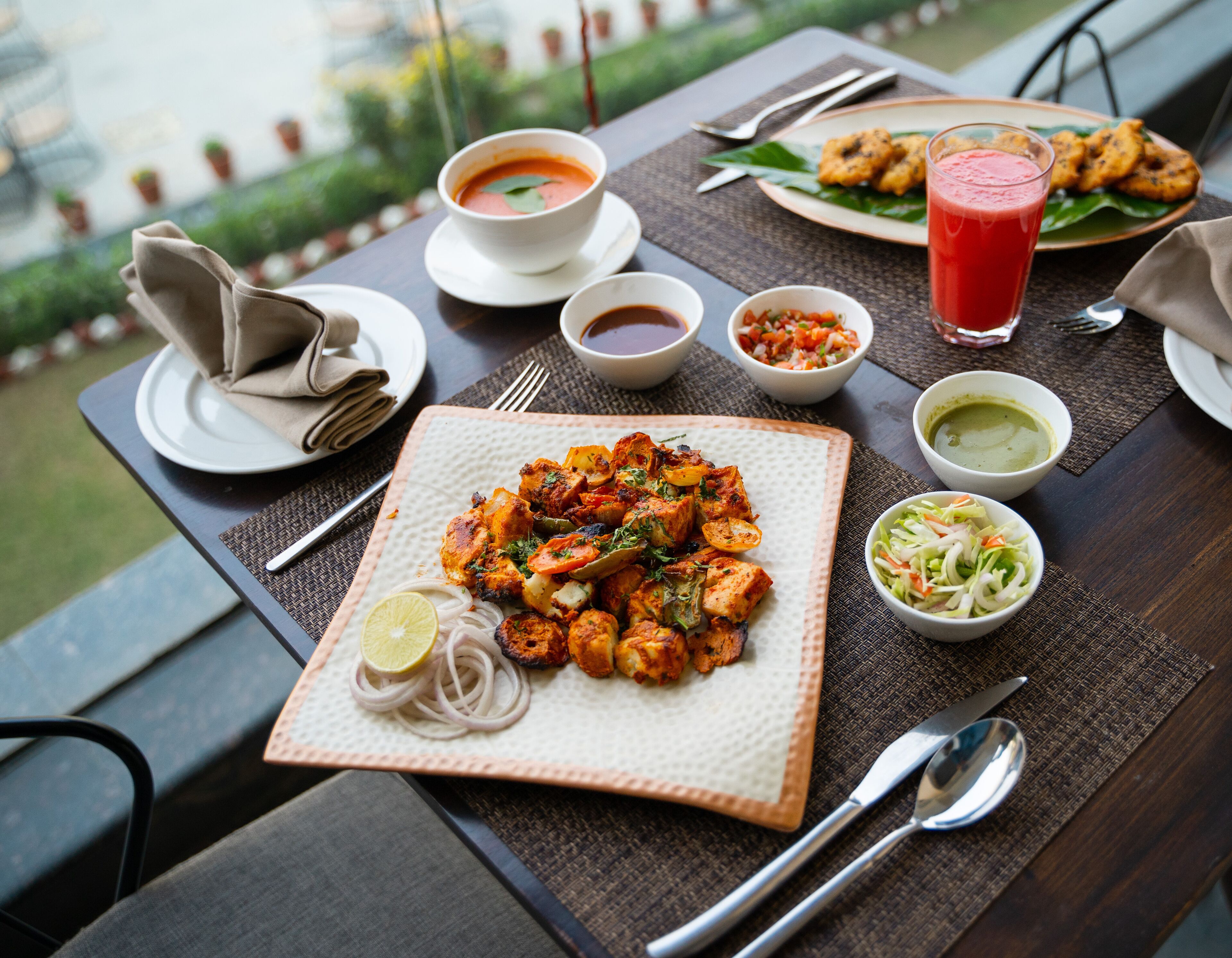 A table set with a meal including grilled chicken, vegetables, and a glass of red drink, with various condiments and a napkin on a woven placemat.
