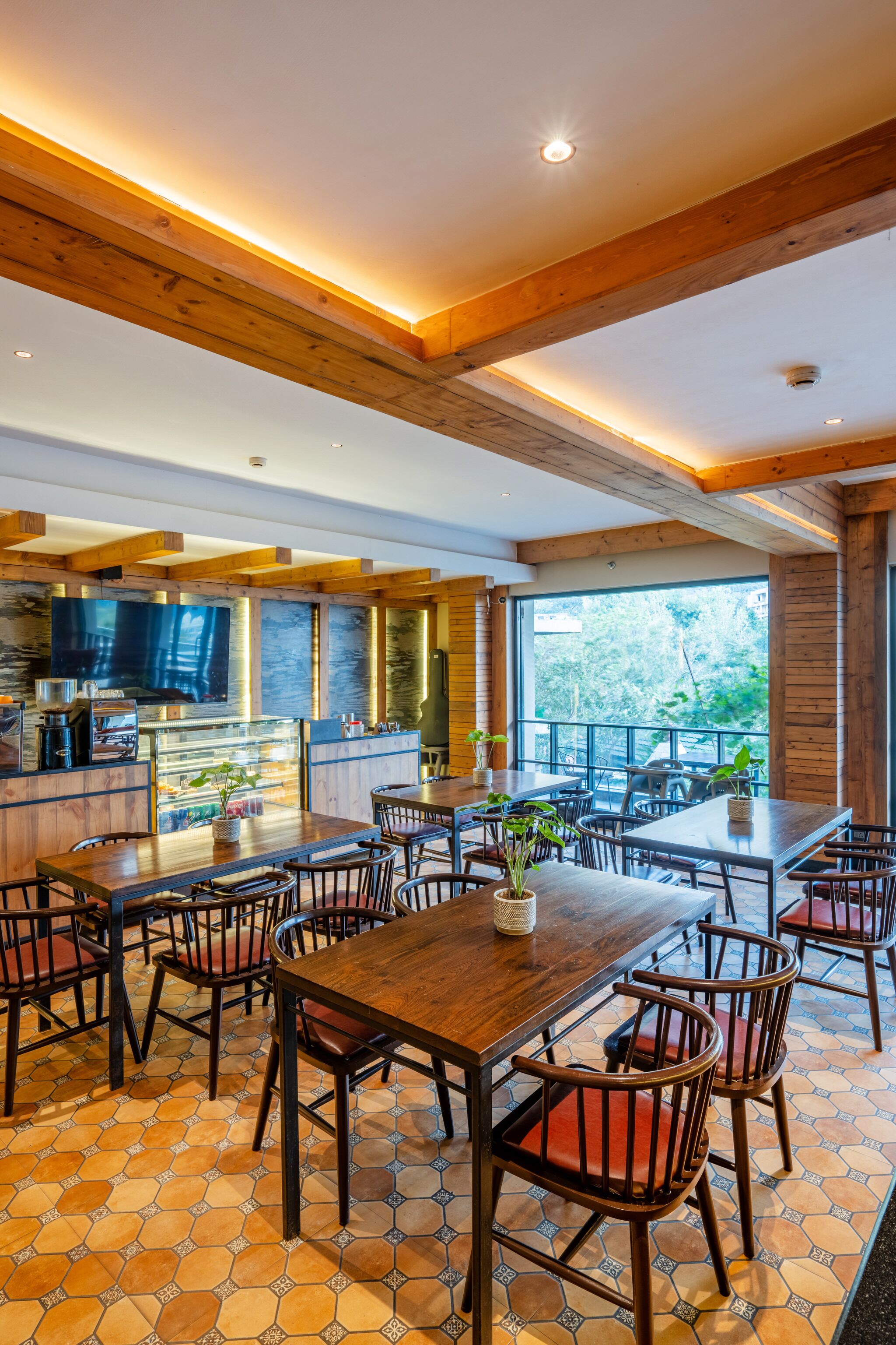 Un comedor amplio con mesas y sillas de madera, una zona de cocina con un televisor grande y vistas a árboles a través de amplias ventanas.