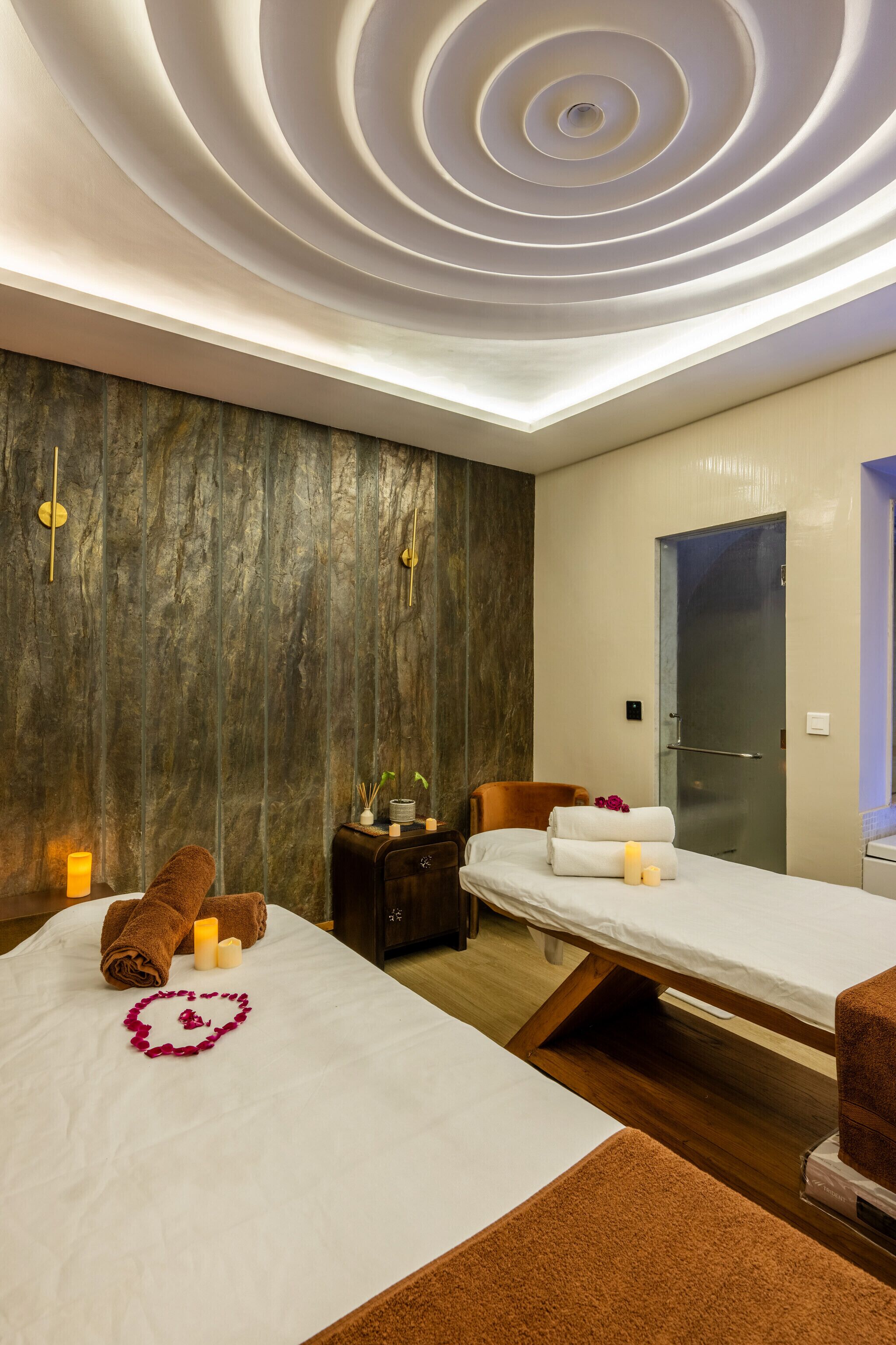 A modern spa room with a wooden wall, a massage table, a massage chair, and a ceiling with circular light fixtures.