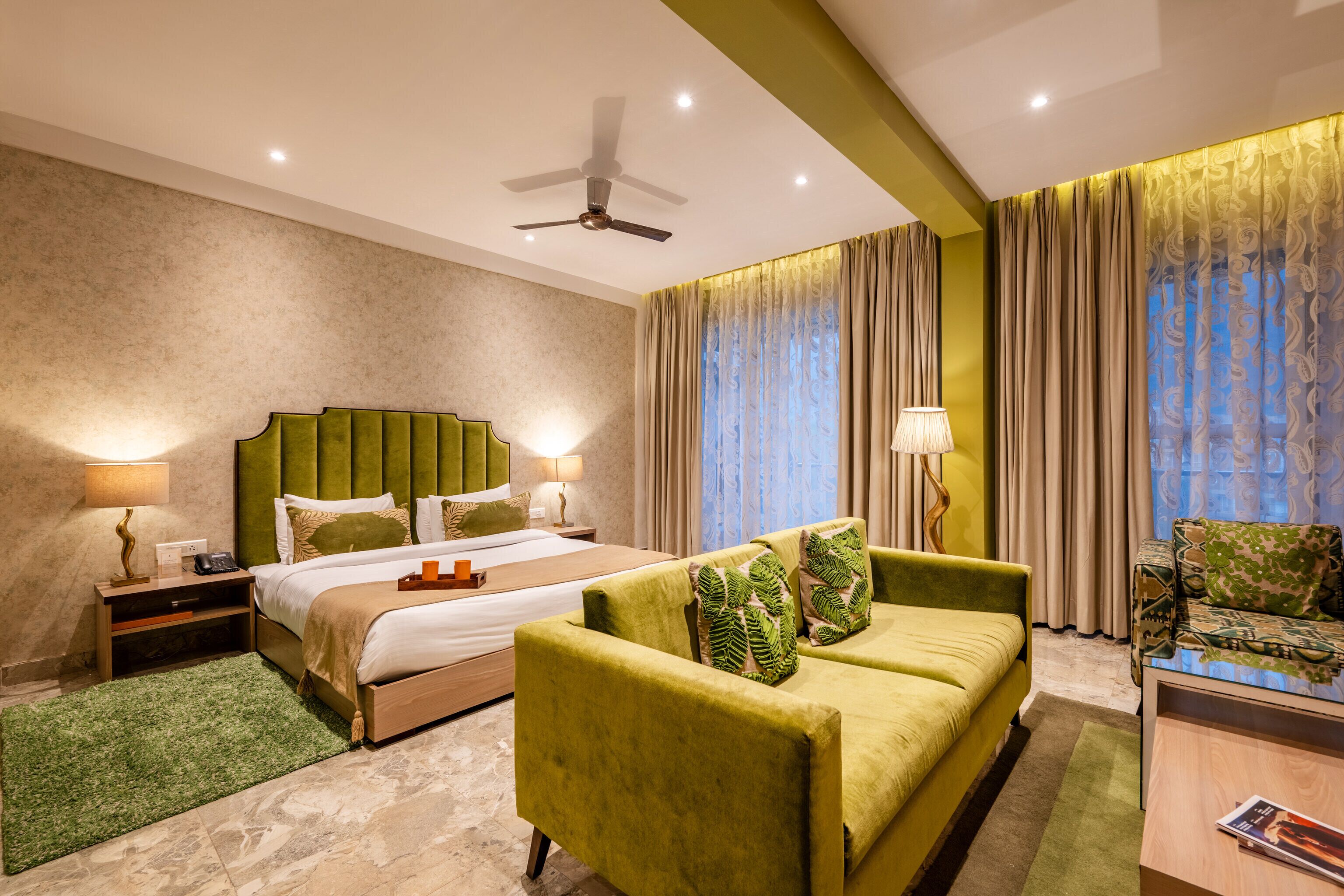 Una habitación de hotel con una cama, un sillón verde, un sofá, una mesita de noche, una lámpara y un ventilador de techo.