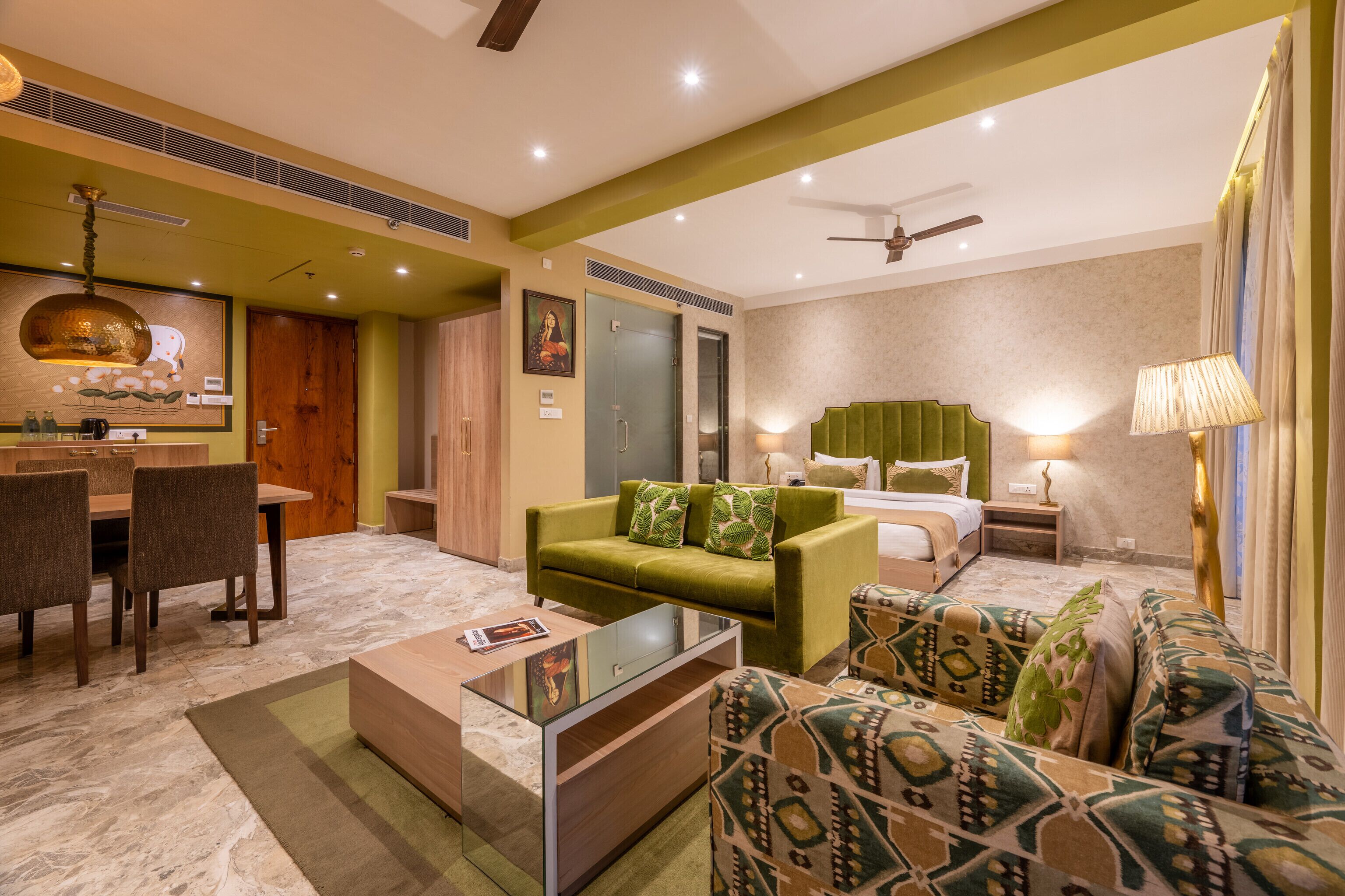 Quarto de hotel com sofá verde, poltrona estampada, área de jantar com mesa e cadeiras, cama com cabeceira verde e ventilador de teto.