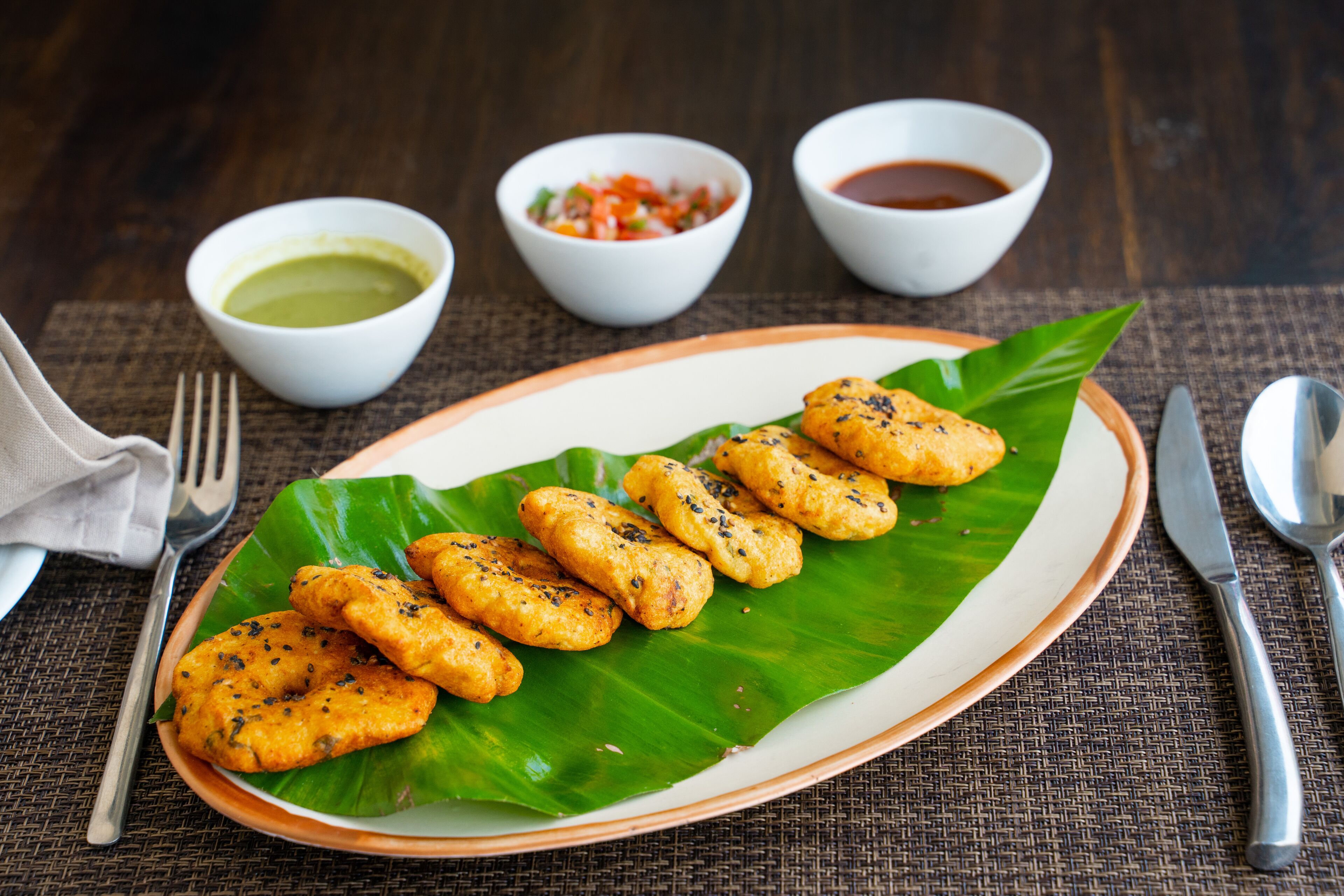 A plate of fried snacks on a banana leaf with three small bowls of sauces.