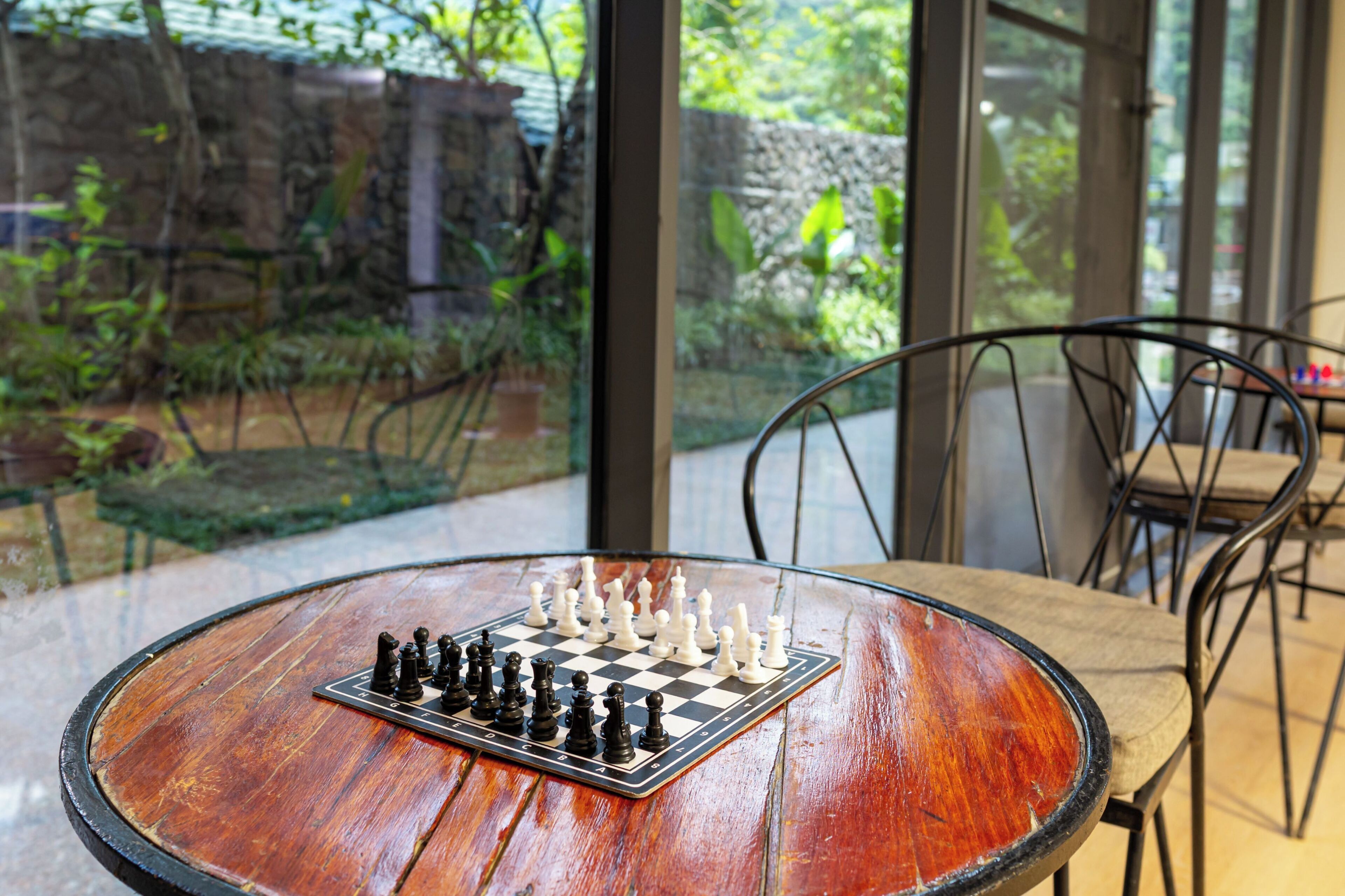A chessboard on a round wooden table with a view of a garden through a glass door.