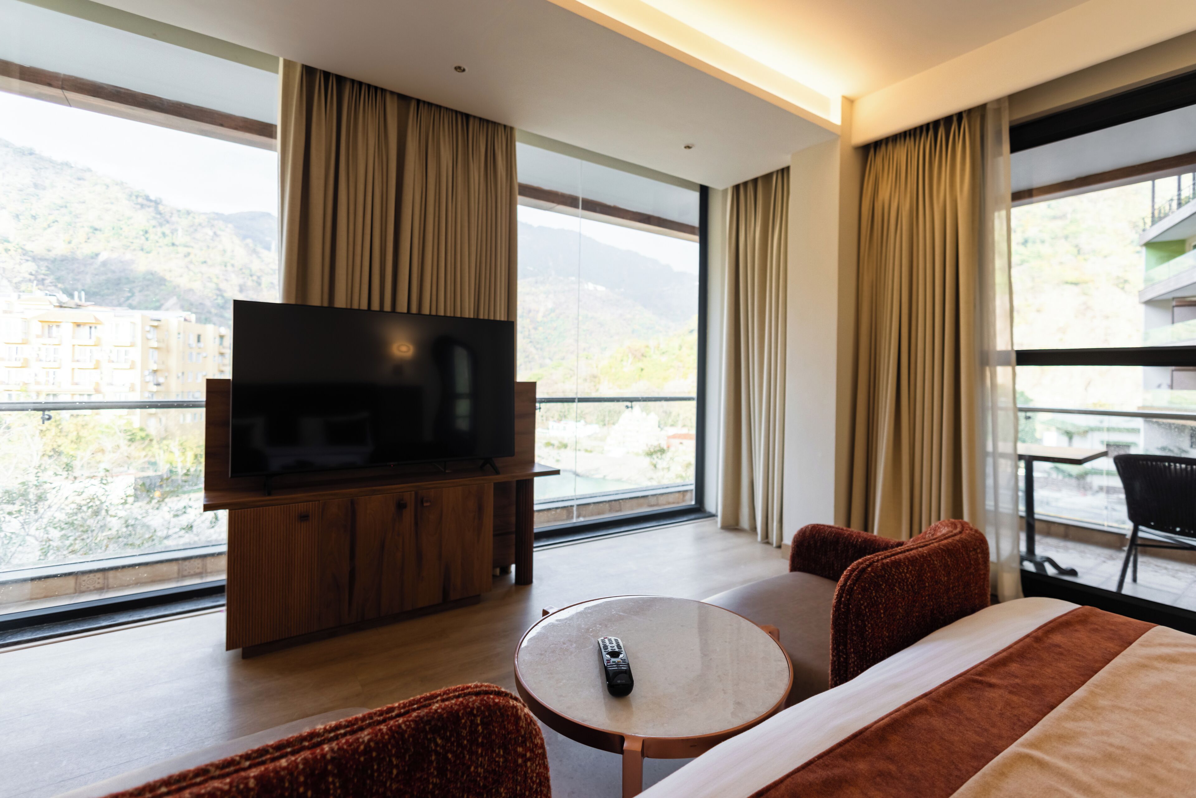 Hotelkamer met een groot raam, een flatscreen televisie op een houten kast, een ronde tafel, twee fauteuils en een bed met een gestreepte deken.