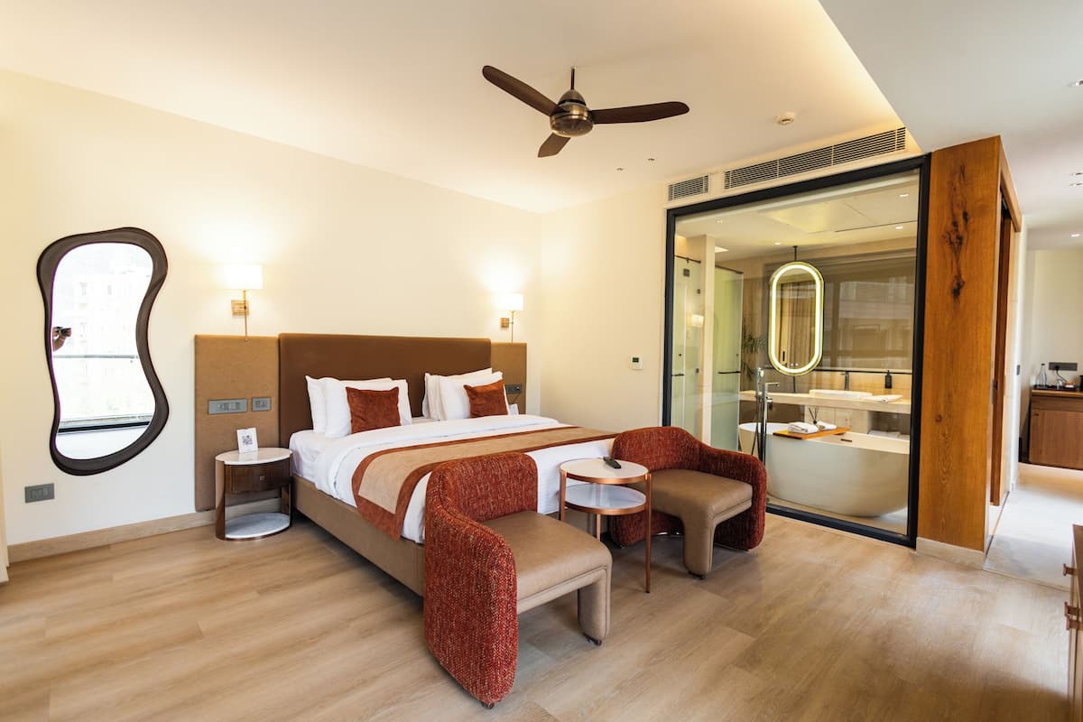 A modern hotel room with a large bed, a sitting area with a table and chairs, a ceiling fan, and a glass-enclosed bathroom.