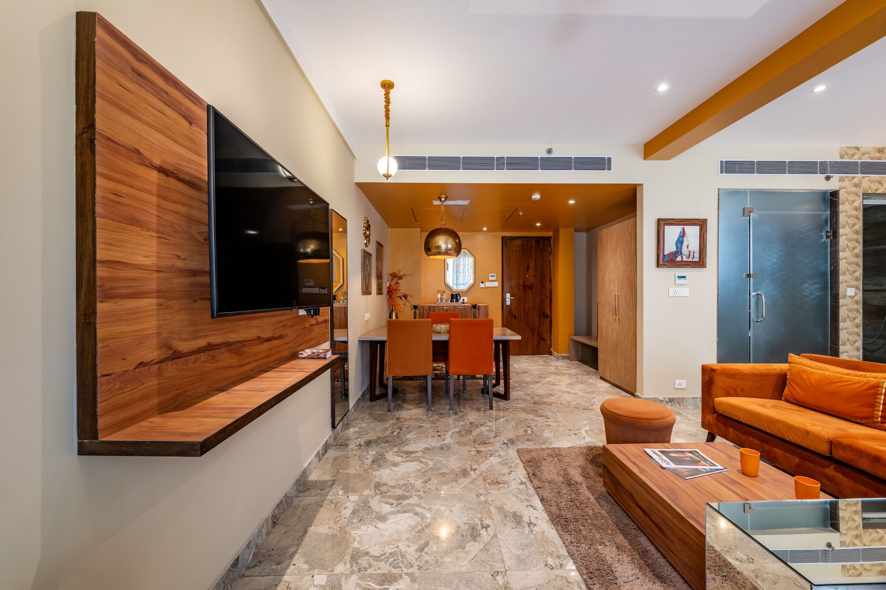 A modern living room with a flat-screen TV, wooden accent wall, orange sofa, and a dining area with a table and chairs.
