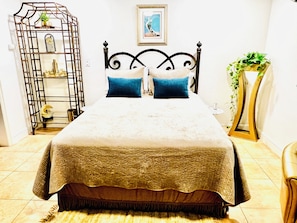 A bed with a headboard, a bedside table, a wall clock, a framed picture, and a decorative plant.
