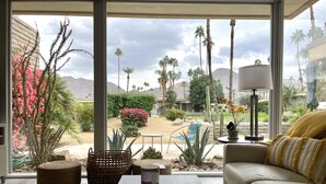 Un soggiorno moderno con una grande finestra, un divano, un tavolino da caffè e vista su un giardino curato con cactus e palme.
