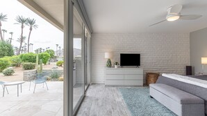 Un soggiorno moderno con una televisione a schermo piatto, una parete in mattoni bianchi e una porta scorrevole in vetro che dà su un patio esterno con sedie e un cactus.