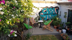 Een terras met een muurschildering, groene stoelen, een tafeltje en verschillende potplanten.