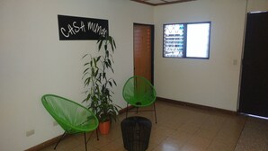 Een kamer met een betegelde vloer, een potplant, twee groene stoelen, een zwarte mand, een houten deur, een raam met een rasterpatroon en een zwart bord aan de muur.