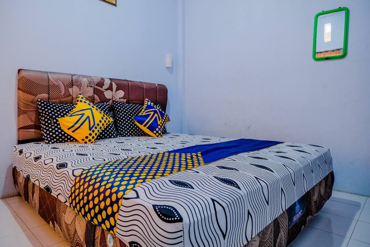 A neatly made bed with patterned bedding and pillows, a headboard, and a small green-framed mirror on the wall.