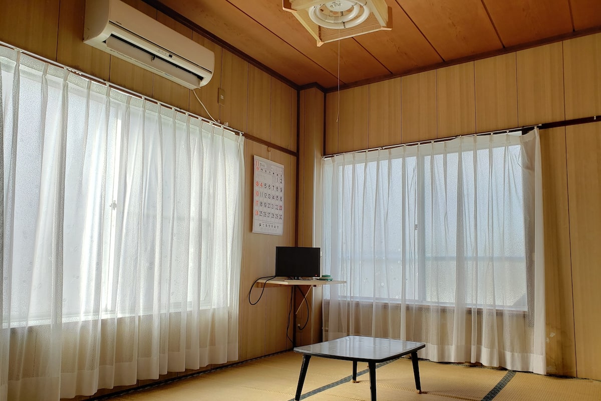 A room with tatami mat flooring, a small table, a TV on a stand, and sheer curtains.