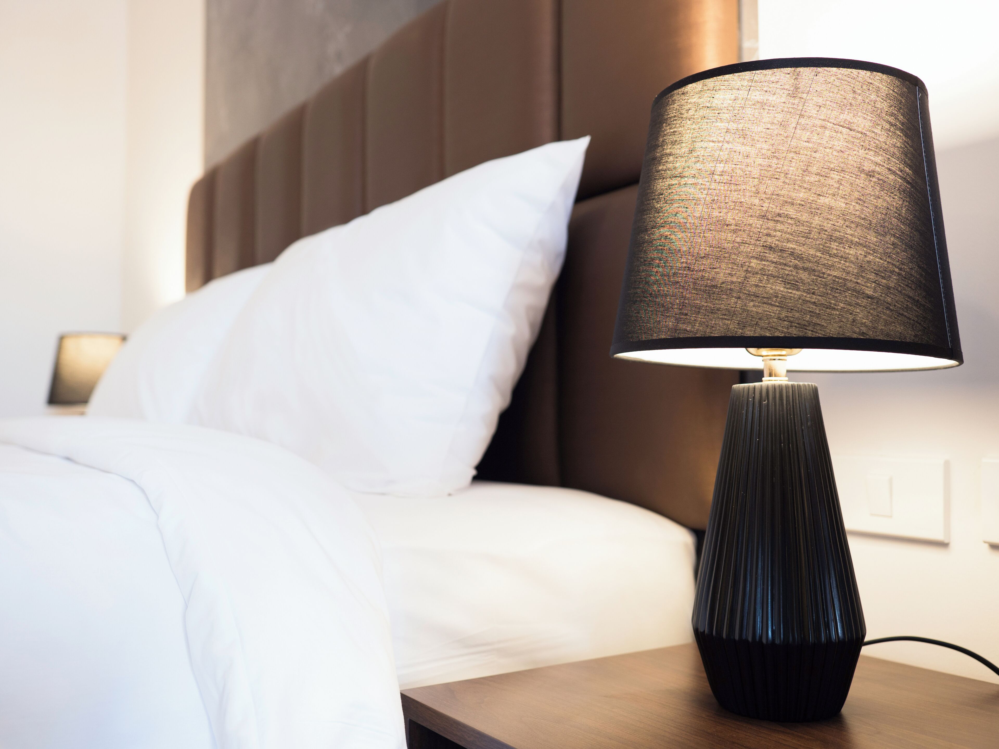 Ein ordentlich bezogenes Bett mit weißen Leinen, eine schwarze, strukturierte Lampe auf einem hölzernen Nachttisch und ein braunes Kopfteil.