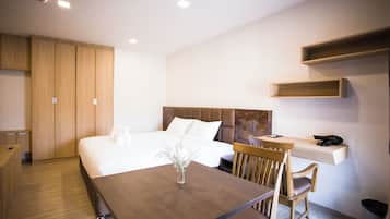 Una habitación de hotel moderna con una cama grande, una mesa de comedor de madera, una silla, un pequeño florero con flores y un escritorio con un teléfono y una bolsa negra.
