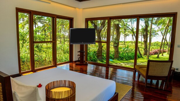 A bedroom with a large bed, a television, and a view of a tropical garden through large windows.