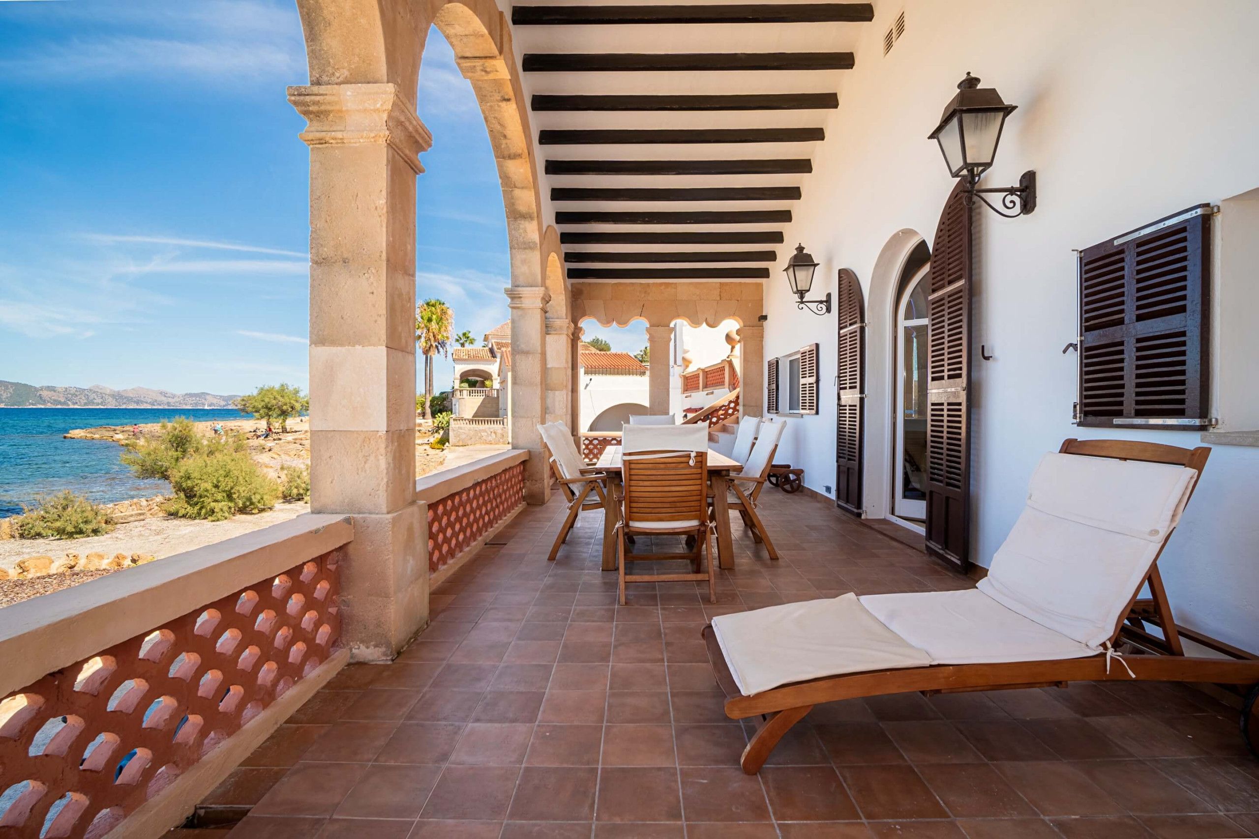 A covered outdoor seating area with a view of the sea, featuring a table and chairs, and a lounge chair.