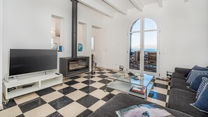 A living room with a black and white checkered floor, a large flat-screen TV, a fireplace, and a view of the sea through a glass door.