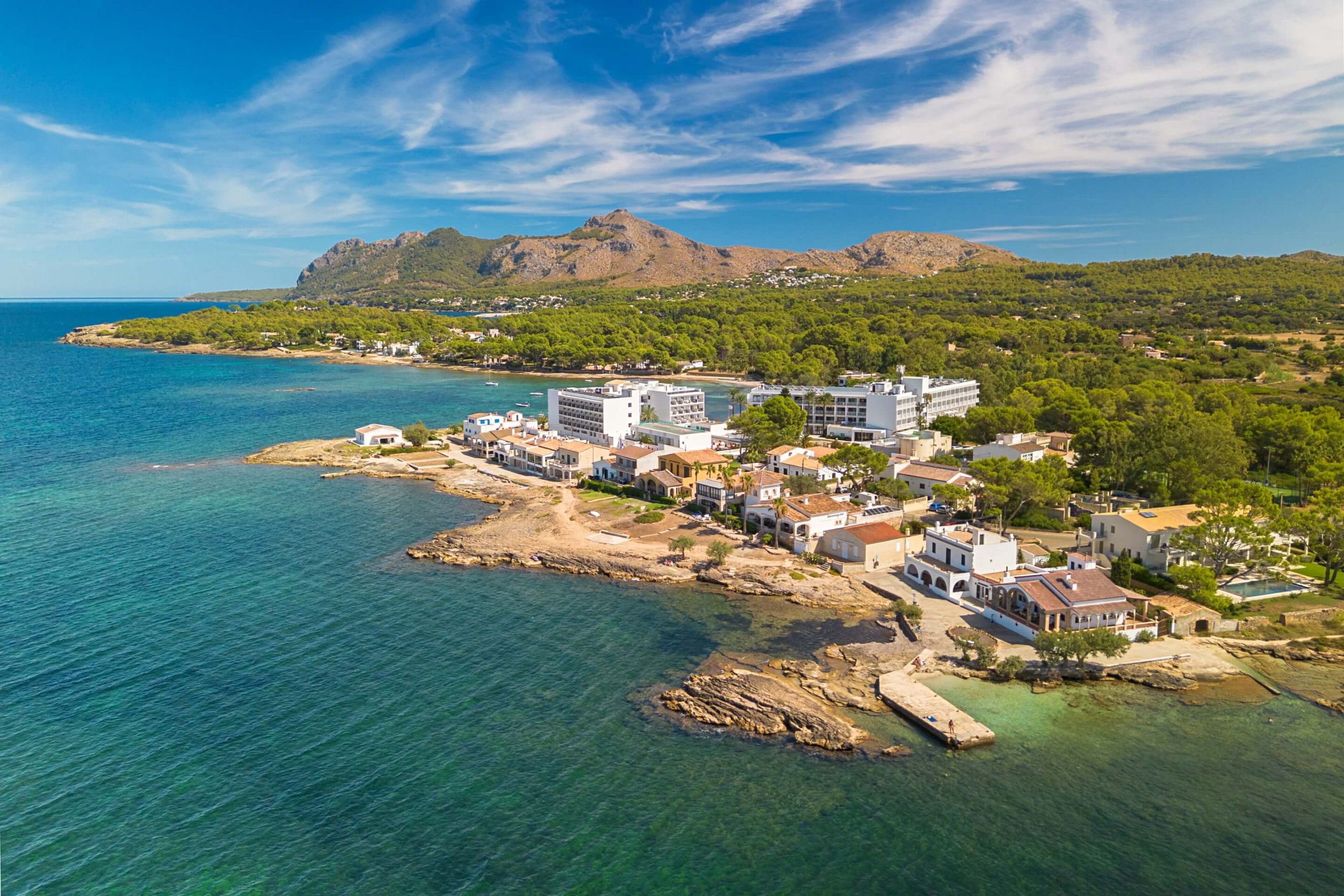 Un pueblo costero con edificios a lo largo de la orilla, una playa rocosa y una montaña al fondo.