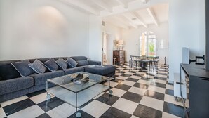 A spacious living room with a checkered floor, a dark grey sofa with patterned cushions, a glass coffee table, and a dining area with a table and chairs.