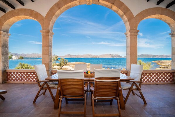 An outdoor seating area with two wooden tables and chairs, overlooking a coastal landscape with clear blue waters and distant hills.
