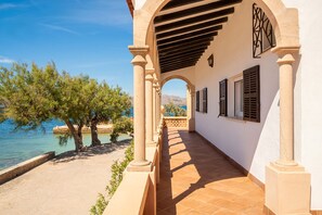 A corridor with columns, a view of the sea, and a building with shutters.