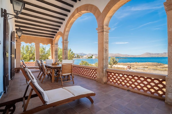 A covered outdoor seating area with a table and chairs, overlooking a body of water and a mountainous landscape. - Villa Es Clot sea view 8 people BBQ (Alcúdia)