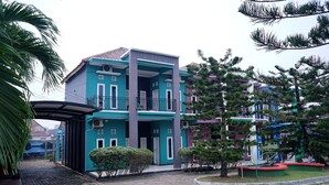 Un edificio de varios pisos con fachada azul y blanca, un acceso cubierto y un patio empedrado con árboles y arbustos pequeños.
